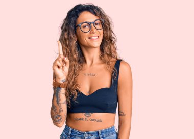 Gündelik giysiler ve gözlük takan Latin kökenli genç bir kadın bir numaralı parmağı gösterip işaret ederken kendinden emin ve mutlu bir şekilde gülümsüyor.. 