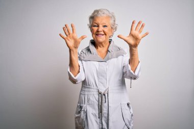 Son sınıf güzeli, gri saçlı, sıradan bir ceket giyen beyaz arka plan üzerinde duran ve parmaklarını on numarayı gösteren, kendinden emin ve mutlu bir şekilde gülümseyen bir kadın..