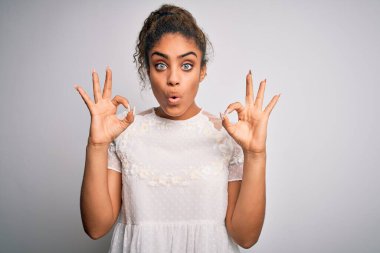 Beyaz arka plan üzerinde sıradan tişörtler giyen Afro-Amerikan genç ve güzel bir kız. Şaşırmış ve şaşırmış görünüyor. Tamam sembolünü parmaklarıyla yapıyor. Çılgın ifade