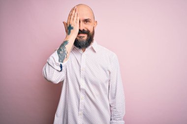 Sakallı yakışıklı, zarif gömlekli, soyutlanmış pembe arka planda bir gözünü kapayan, yüzünde kendine güvenen bir gülümseme ve sürpriz bir duygu olan kel bir adam..