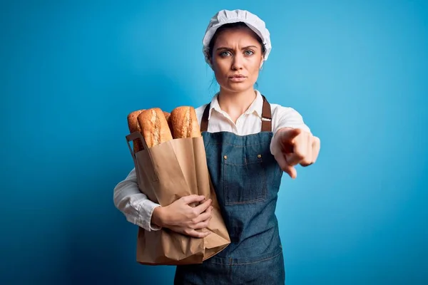 年轻美丽的面包师女人 蓝眼睛 手握纸袋 手拿着面包 手指指向相机 正面是积极自信的手势 — 图库照片