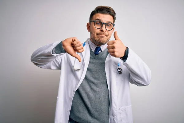 Young doctor man with blue eyes wearing medical coat and stethoscope over isolated background Doing thumbs up and down, disagreement and agreement expression. Crazy conflict