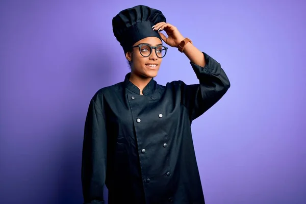 Young african american chef girl wearing cooker uniform and hat over purple background smiling confident touching hair with hand up gesture, posing attractive and fashionable