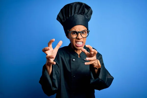 Young african american chef woman wearing cooker uniform and hat over blue background Shouting frustrated with rage, hands trying to strangle, yelling mad