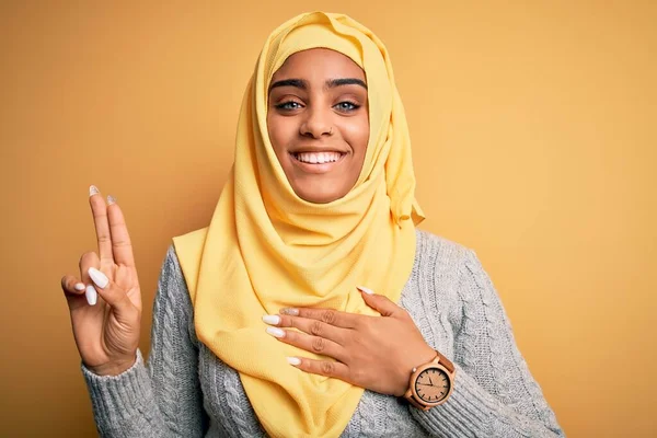 Young beautiful african american girl wearing muslim hijab over isolated yellow background smiling swearing with hand on chest and fingers up, making a loyalty promise oath