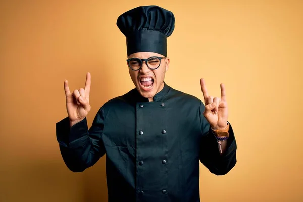 Young brazilian chef man wearing cooker uniform and hat over isolated yellow background shouting with crazy expression doing rock symbol with hands up. Music star. Heavy music concept.
