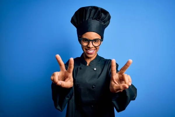 Young african american chef woman wearing cooker uniform and hat over blue background smiling with tongue out showing fingers of both hands doing victory sign. Number two.