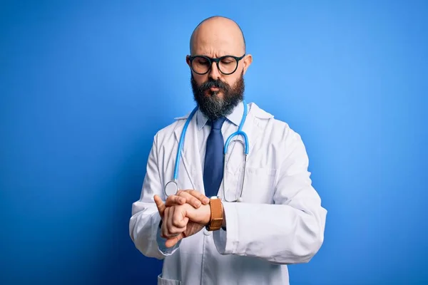 英俊的秃头医生 留着胡子 戴着眼镜 带着听诊器 带着蓝色背景检查腕表的时间 放松而自信 — 图库照片