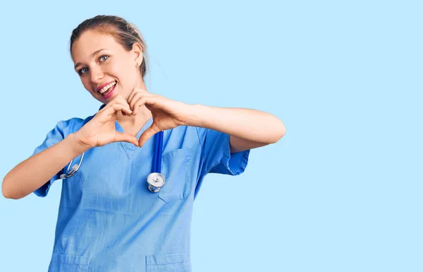 医師の制服を着た若い美しいブロンドの女性と手でハートシンボルの形をして愛に笑みを浮かべて聴診器 ロマンチックなコンセプト — ストック写真