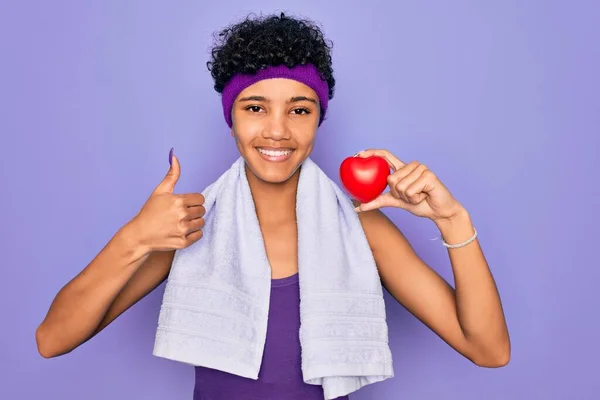 Beautiful african american afro sporty woman doing exercise wearing towel holding heart happy with big smile doing ok sign, thumb up with fingers, excellent sign