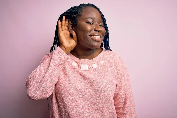 African american plus size woman with braids wearing casual sweater over pink background smiling with hand over ear listening an hearing to rumor or gossip. Deafness concept.