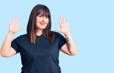 Genç ve cüsseli kadın gündelik kıyafetler giyer ve parmaklarını on numarayla gösterir ve gösterirken kendinden emin ve mutlu gülümser.. 