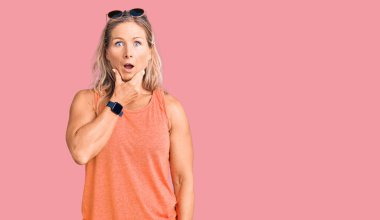 Orta yaşlı, yaz kıyafetleri ve güneş gözlüğü takan sarışın bir kadın. İnançsızlıktan, şaşkınlıktan ve elleri çenesinde şaşkınlıktan büyülenmiş görünüyor. 