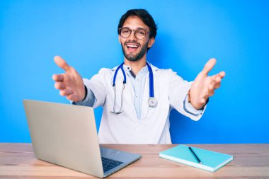 Klinikte çalışan yakışıklı, doktor üniforması giyen, kucaklaşmak için kollarını açmış kameraya bakan bir İspanyol. Mutluluğu kucaklayan neşeli bir ifade.. 