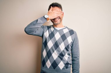 İzole edilmiş beyaz arka planda dikilen sıradan süveter giyen genç yakışıklı bir adam. Gülümsüyor ve gülümsüyor. Yüzünde şaşkınlık için gözleri örtüyor. Kör kavram.