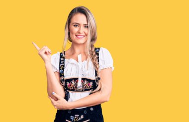 Oktoberfest elbisesi giyen, yüzünde kocaman bir gülümseme olan, eli ve parmağı kameraya bakan genç, güzel sarışın kadın.. 