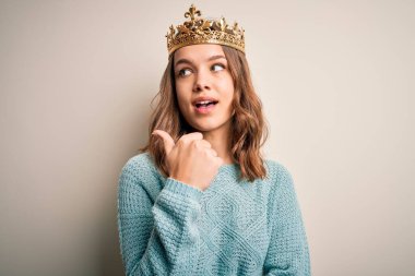 Soyutlanmış arka planda kraliçe tacı takan genç sarışın kız gülümsüyor ve başparmağıyla yan tarafa bakıyor..
