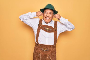 Sarı arka planda Alman geleneksel ahşap şenliği kıyafeti giyen yaşlı gri saçlı adam parmaklarıyla kulaklarını çekerek gülümsüyor, komik bir jest yapıyor. Seçme sorunu