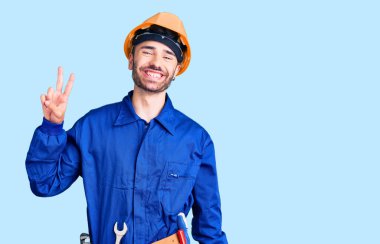 İşçi üniforması giyen genç İspanyol adam iki numarayı gösterip işaret ederken kendinden emin ve mutlu bir şekilde gülümsüyor.. 