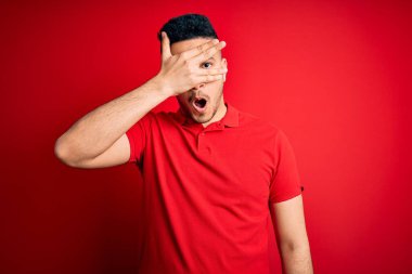 İzole edilmiş arka planda kırmızı polo giyen genç yakışıklı adam şok içinde yüzünü ve gözlerini kapayarak utangaç bir ifadeyle parmaklarının arasından bakıyor..