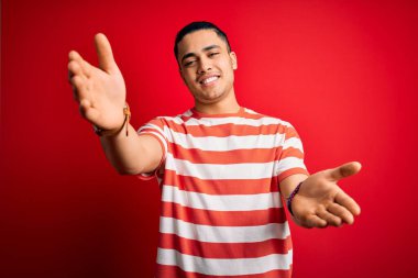 Sıradan çizgili tişört giyen genç Brezilyalı adam izole edilmiş kırmızı arka planda dikilip sarılmak için kollarını açıp kameraya gülümsüyor. Mutluluğu kucaklayan neşeli bir ifade..