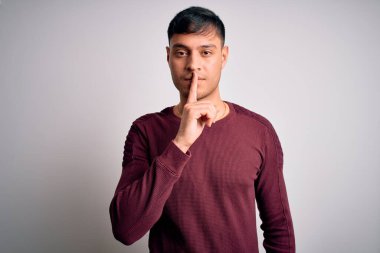 Beyaz izole edilmiş arka plan üzerinde dikilmiş, dudakları parmaklarıyla sessiz olmak isteyen, gündelik tişört giyen genç, yakışıklı İspanyol adam. Sessizlik ve gizli konsept.