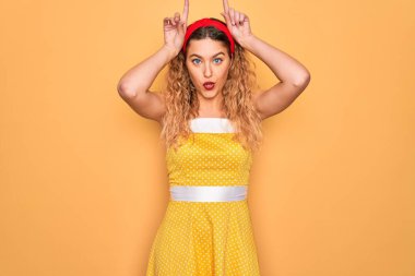 Sarı arka planda dikilen mavi gözlü güzel sarışın kadın boynuzları gibi parmağıyla komik hareketler yapıyor.