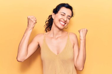 Genç ve güzel İspanyol bir kadın gündelik kıyafetler giyip çok mutlu ve heyecanlı bir şekilde kolları havada, gülümseyen ve başarı için bağıran kazanan bir jest yapıyor. kutlama konsepti. 