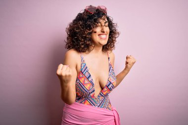 Pembe arka planda mayo ve güneş gözlüğü takan genç, güzel bir Arap kadın kolları havada, gülümseyen ve başarı için bağıran mutlu ve heyecanlı bir jest yapıyor. Kutlama konsepti.