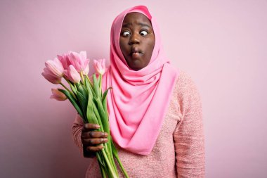 Genç, Afro-Amerikan, iri yarı, Müslüman bir kadın elinde pembe lale buketi, dudaklarıyla balık suratı, çılgınca ve komik bir jest yapıyor. Komik ifade.