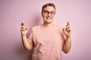Genç, kızıl saçlı, günlük tişört giyen izole edilmiş pembe arka plan üzerinde duran umut ve gözler kapalı gülümseyerek el hareketi yapan yakışıklı bir adam. Şans ve batıl inanç..