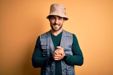 Tatilde sakallı yakışıklı turist sarı arka planda kaşif şapkası takıyor ve parmaklarını çapraz yapıp gevşemiş ve neşeli gülümsüyor. Başarı ve iyimser