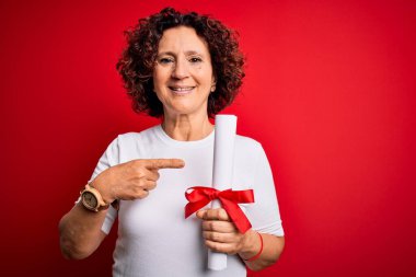 Orta yaşlı kıvırcık saçlı, elinde üniversite diploması olan bir kadın elini ve parmağını göstererek mutlu bir şekilde mezun oldu.