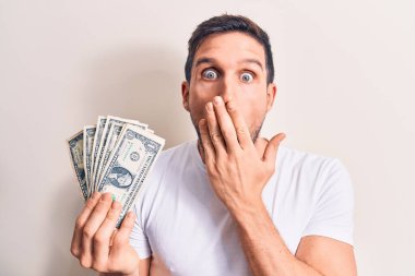 Genç yakışıklı bir adam, izole edilmiş beyaz arka planda bir deste banknot tutuyor. Ağzı elle kaplıyor, şok geçiriyor ve hata yapmaktan korkuyor. Şaşırtıcı ifade.