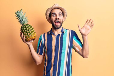 Sakallı, yaz şapkalı ve tişörtlü genç yakışıklı adam ananas tutarak başarıyı kutluyor mutlu bir gülümsemeyle ve kazanan yüz ifadesiyle elini kaldırarak. 