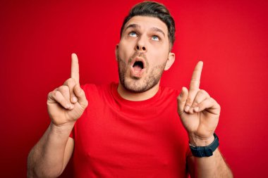 Mavi gözlü genç adam, kırmızı izole edilmiş arka plan üzerine gündelik tişört giyiyor. Şaşırmış ve şaşırmış bir şekilde yukarı bakıyor, parmaklarıyla işaret ediyor ve kollarını kaldırıyordu..