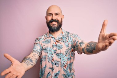 Sakallı, dövmeli, pembe arka planda sıradan bir çiçek gömleği giyen yakışıklı kel adam sarılmak için kollarını açmış gülümseyen kameraya bakıyor. Mutluluğu kucaklayan neşeli bir ifade..