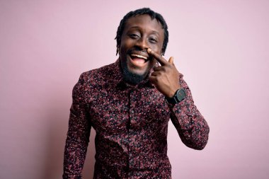 Genç, yakışıklı, Afro-Amerikan bir adam pembe arka planda dikilmiş, parmaklarıyla yüzünü ve burnunu göstererek neşeli bir şekilde gülümsüyor. Güzellik konsepti