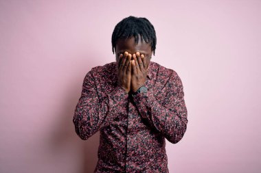 Genç, yakışıklı, Afro-Amerikalı bir adam pembe arka planda dikilmiş ağlarken suratını ellerle kapatan bir pembe tişört giyiyor. Depresyon kavramı.