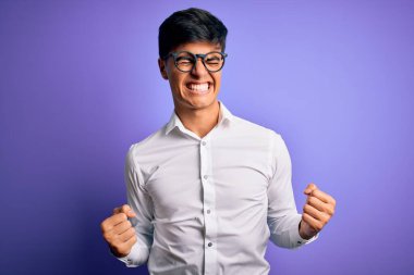 Mor arka planda gömlek ve gözlük takan genç yakışıklı iş adamı mutlu ve heyecanlı bir şekilde kollarını kaldırıp, gülümseyerek ve başarı için bağırarak kazanan bir jest yapıyor. Kutlama konsepti.