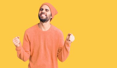 Sakallı, sıradan bir kazak ve yün şapka giyen genç yakışıklı adam çok mutlu ve heyecanlı bir şekilde kolları havada, gülümseyerek ve başarı için çığlık atarak kazanan hareketi yapıyor. kutlama konsepti. 