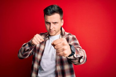 Genç, beyaz, gündelik modern gömlekler giyen, kırmızı arka planda kalan yakışıklı bir adam. Yumruklarını yumrukluyor, saldırıyor, saldırıyor, tehdit ediyor ve şiddet uyguluyor.