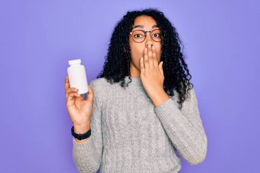 Genç Afro-Amerikalı kıvırcık kadın, elinde haplar tutuyordu. Mor arka planda yalnız duruyordu. Ağzı kapatıyordu.