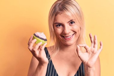 Sarışın, güzel bir kadın, izole edilmiş sarı arka planda tatlı çikolatalı kek yiyor. Tamam işareti yapıyor. Parmaklarıyla, gülümsüyor.