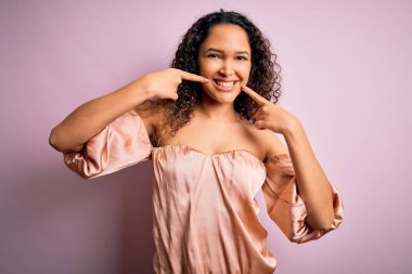 Kıvırcık saçlı, pembe arka planda sıradan tişörtler giyen neşeli bir şekilde gülümseyen ve parmaklarıyla ve ağzıyla işaret eden güzel bir kadın. Diş sağlığı konsepti.