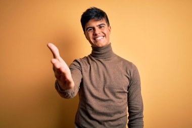 Sarı arka plan üzerine balıkçı yaka kazak giyen yakışıklı genç bir adam selamlama ve hoş geldin demek için gülümseyerek el sıkışır. Başarılı bir iş..