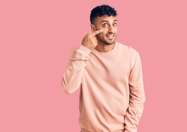 Latin asıllı genç bir adam, gündelik giysiler giyip el ve burnu işaret ediyor, neşeli bir şekilde gülümsüyor. güzellik konsepti 