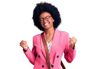 İş kıyafetleri ve deri çanta giyen genç Afro-Amerikalı kadın çok mutlu ve heyecanlı bir şekilde kollarını kaldırıp, gülümseyerek ve başarı çığlıkları atarak zafer jesti yapıyor. kutlama konsepti. 