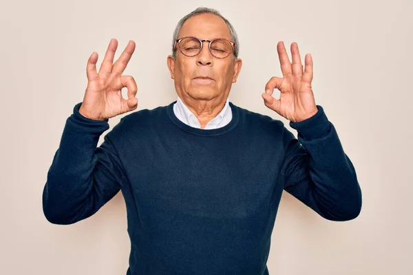 Senior handsome grey-haired man wearing sweater and glasses over isolated white background relax and smiling with eyes closed doing meditation gesture with fingers. Yoga concept.