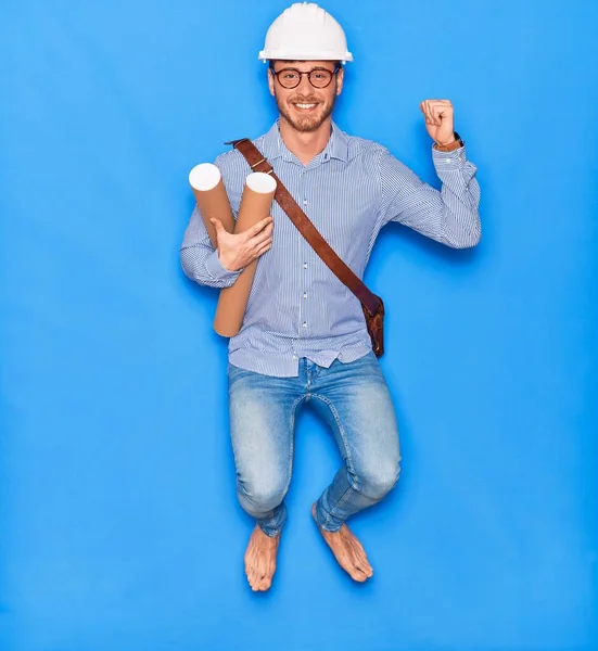 Young handsome architect man wearing leather bag and hardhat smiling happy. Jumping with smile on face holding blueprints celebrating with fist raised over isolated blue background